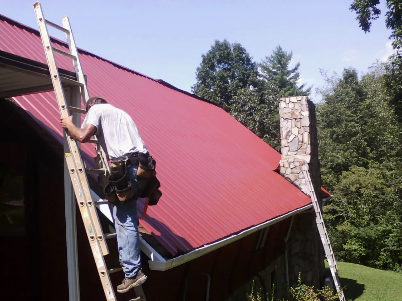 Roofer on a ladder beside a red metal roof and stone chimney — Soffit & Fascia Repair by Roofing Brothers in Keokuk